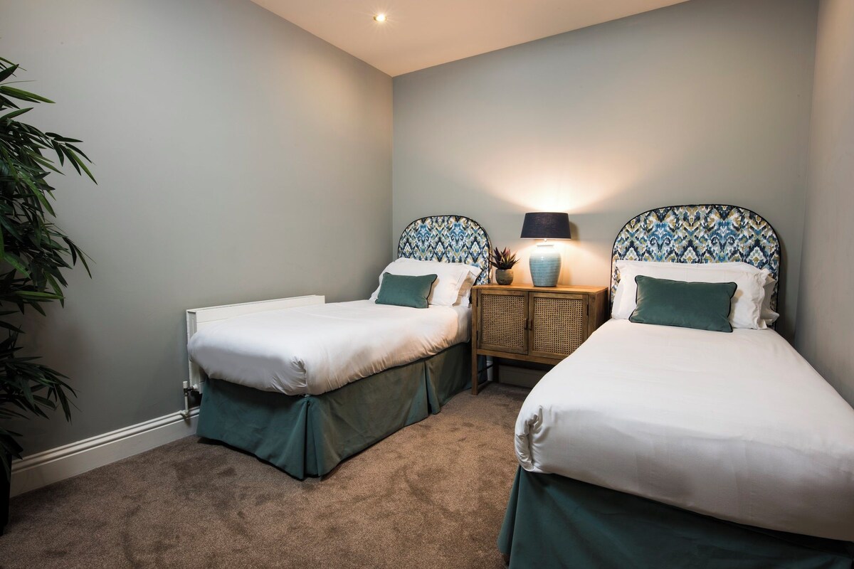 A bedroom with two single beds, a bedside table with a lamp, a small plant, and a large potted plant.