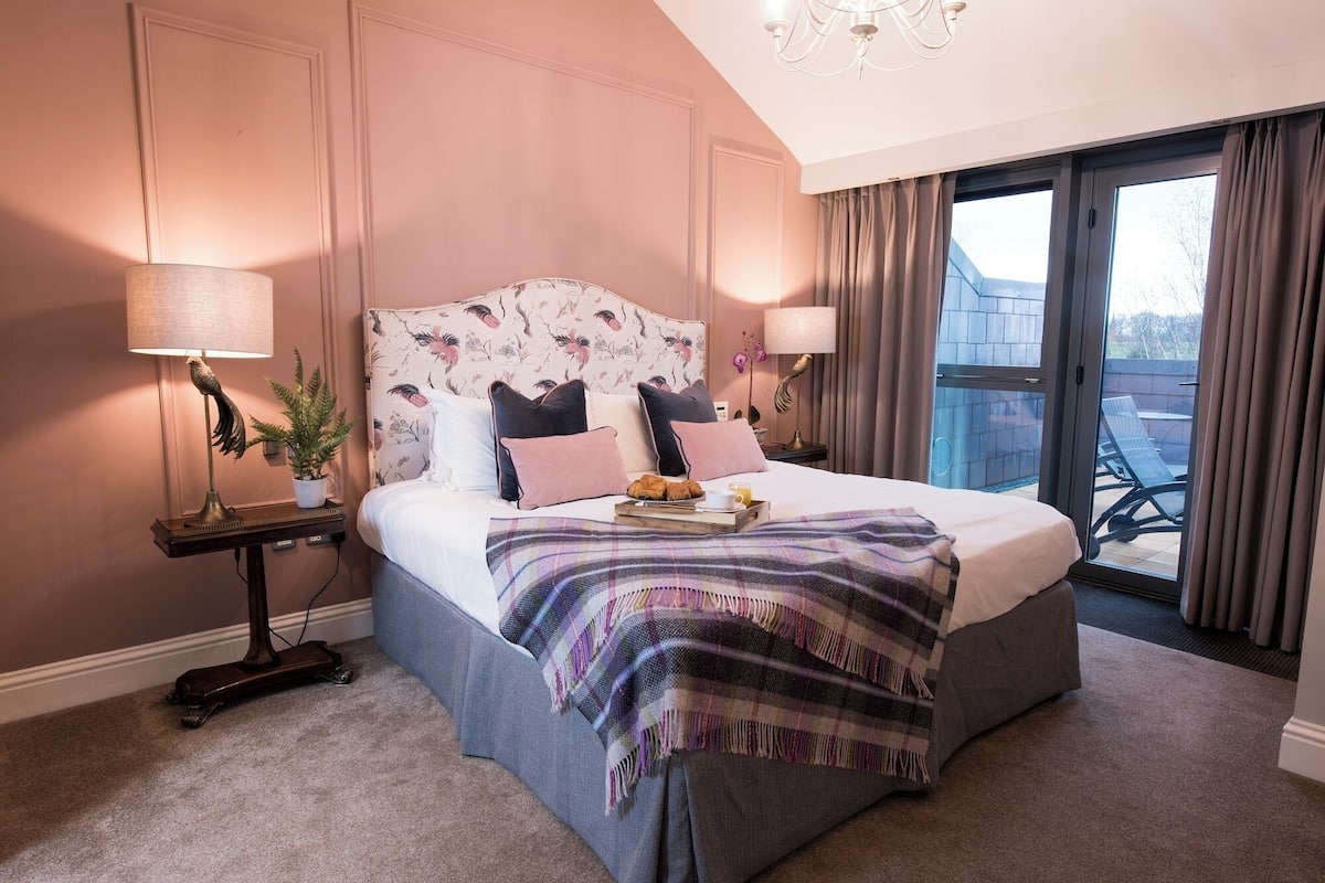 A bedroom with a bed, a nightstand, a lamp, a potted plant, and a view of the outside.