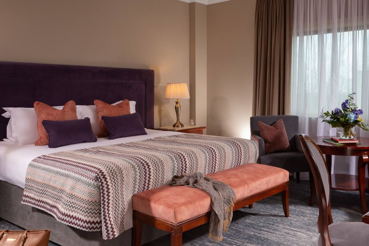 A hotel room with a large bed, a bench, a chair, a table with a vase of flowers, and a lamp.