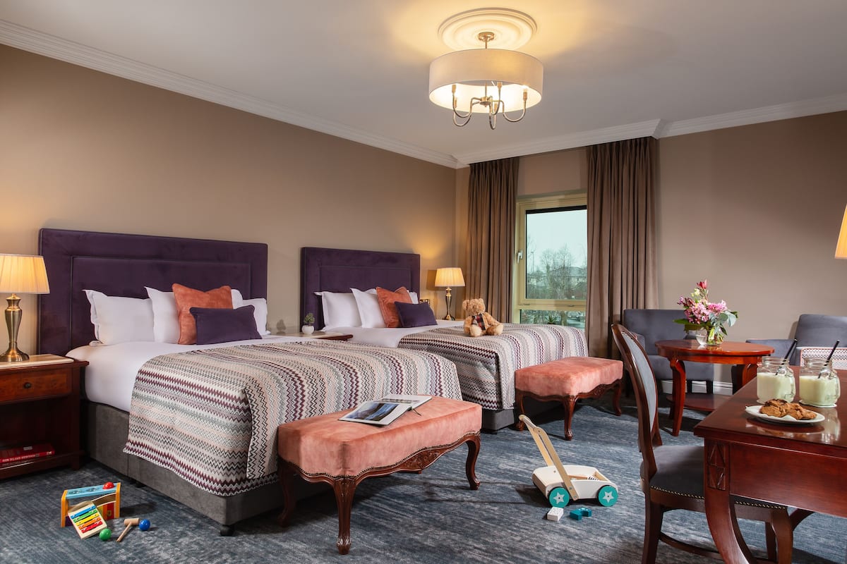 A hotel room with two beds, a dining table, a chair, a small table with a teddy bear, and a window with curtains.