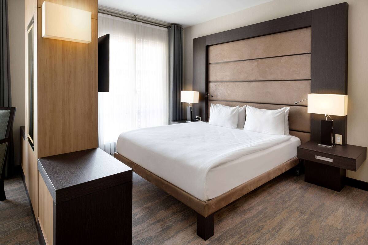 Ein Hotelzimmer mit einem großen Bett, einem Holz-Kopfteil, zwei Nachttischen mit Lampen, einem Flachbildfernseher und einem Schreibtisch mit Stuhl.