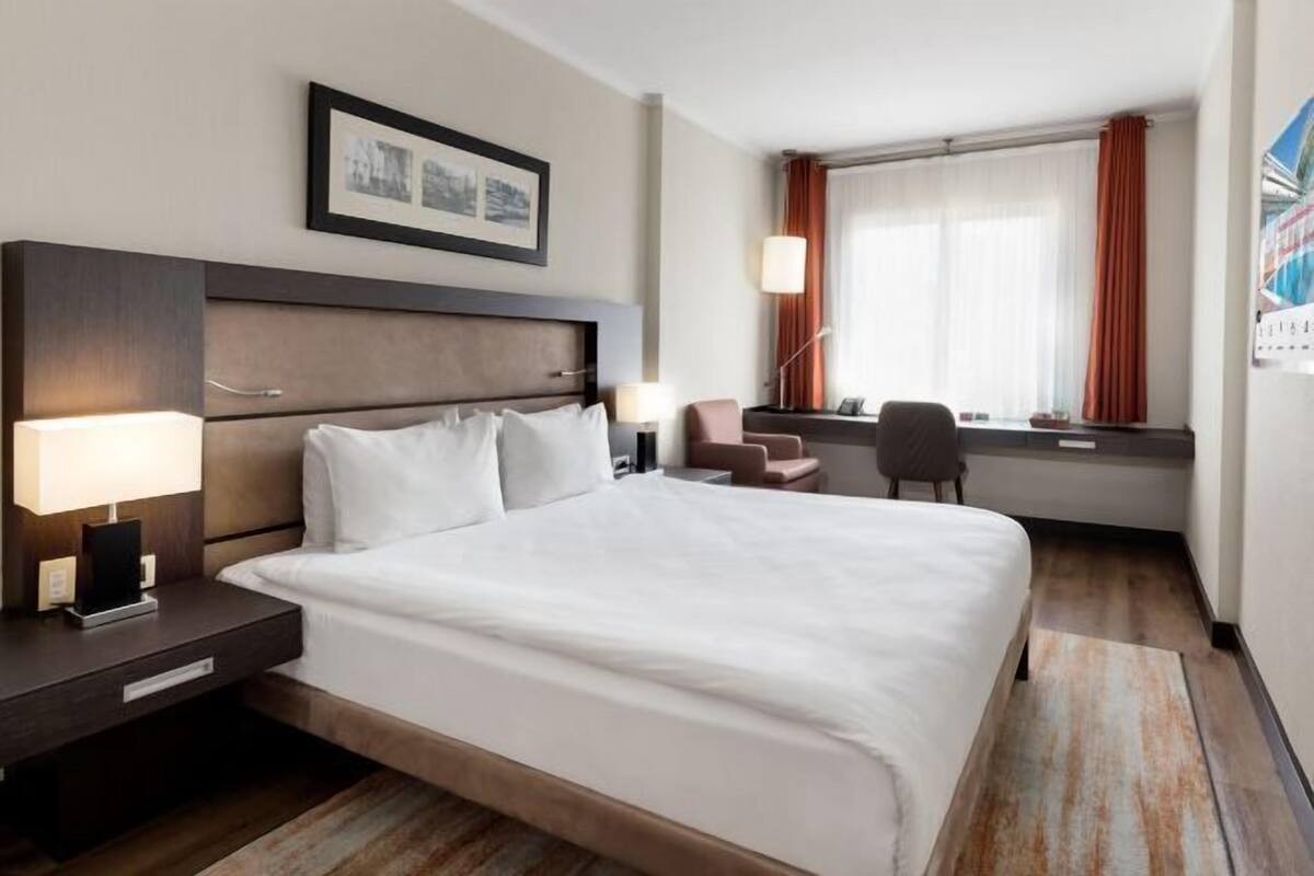 Una habitación de hotel con una cama grande, mesitas de noche, un escritorio y una ventana con cortinas.