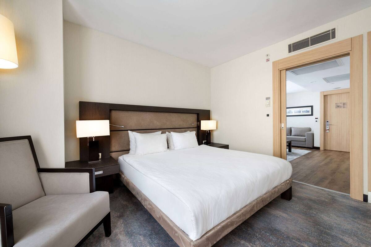 Ein Hotelzimmer mit einem großen Bett, zwei Nachttischen inklusive Lampen, einem Sessel und einer Holztür zu einem weiteren Raum.