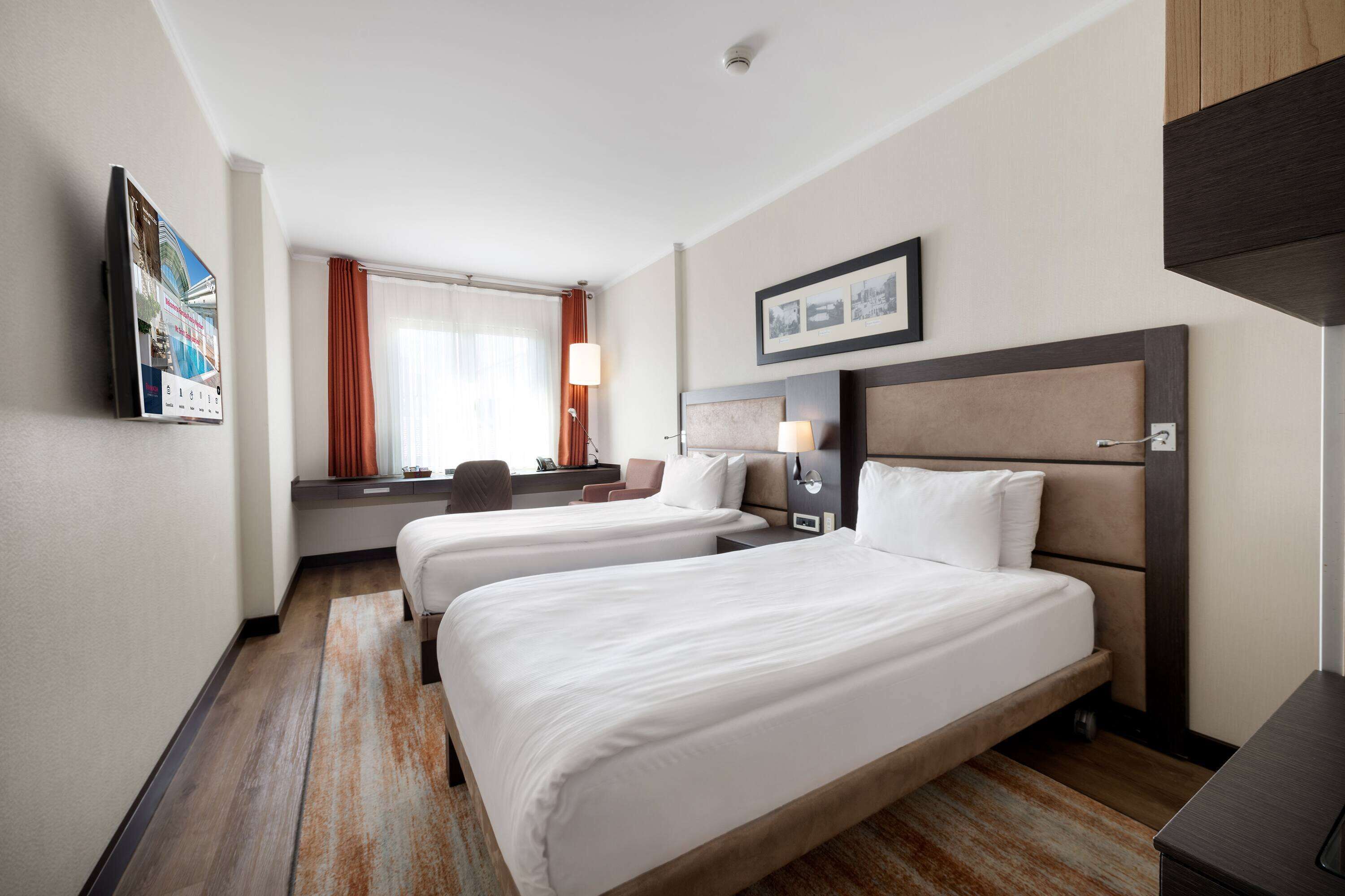 A hotel room with two beds, a desk, a chair, a window with curtains, and a wall-mounted picture.