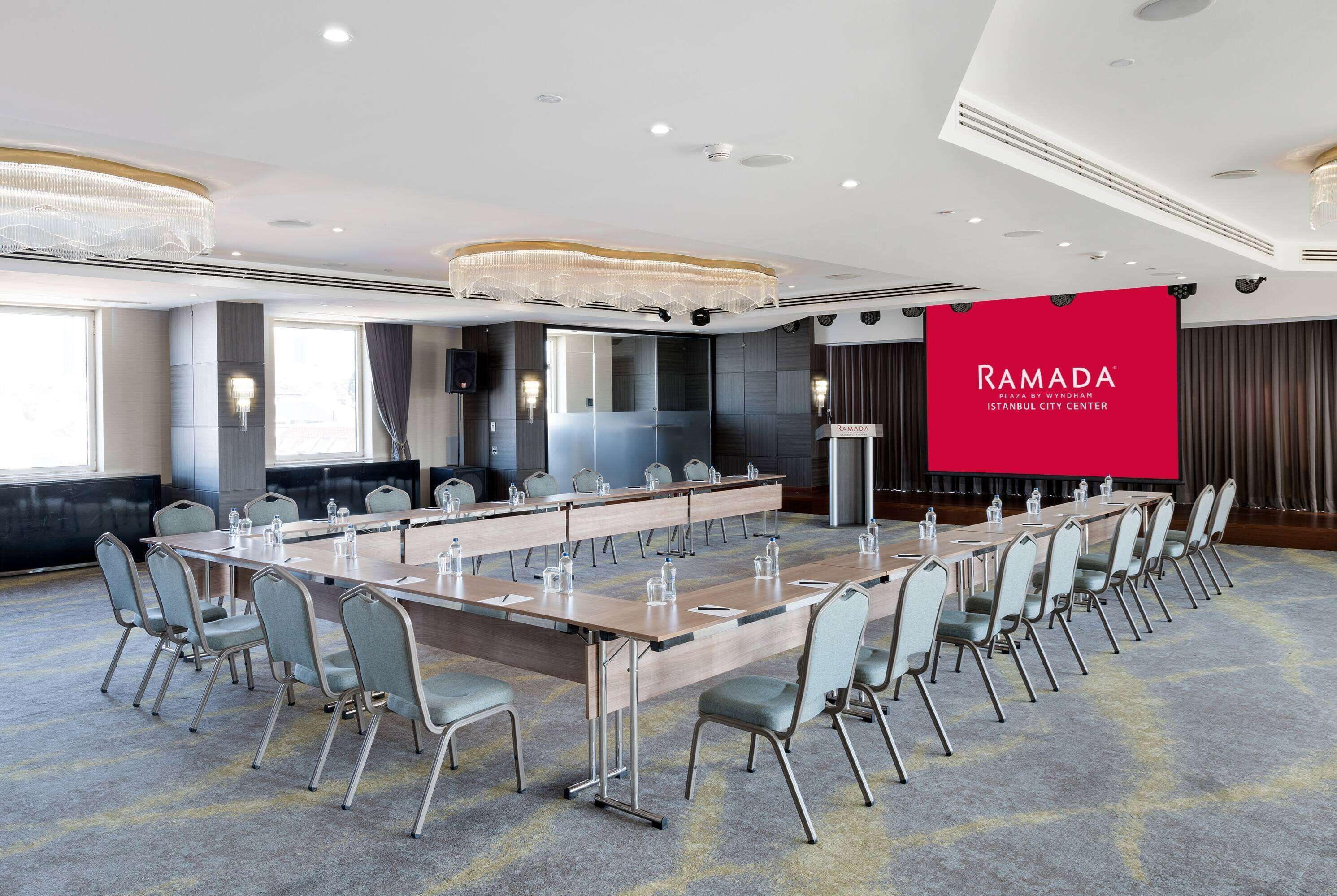 Ein Konferenzraum mit einem langen Tisch, Stühlen und einem großen Bildschirm, der "RAMADA ISTANBUL CITY CENTER" anzeigt.