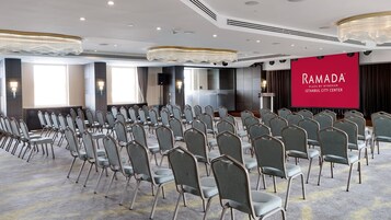Una sala conferenze con file di sedie grigie, un grande schermo che mostra 'RAMADA ISTANBUL CITY CENTER' e un moderno lampadario.