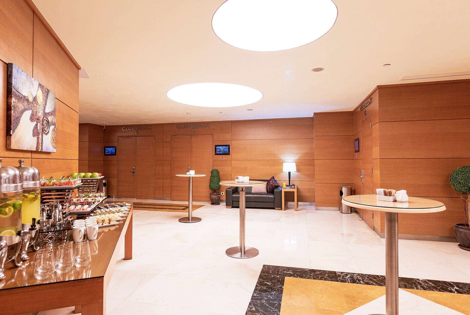 Un hall d’hôtel comprenant un comptoir d’accueil, deux salles de réunion désignées par les lettres A et B, un buffet proposant divers plats et une station de préparation de café.