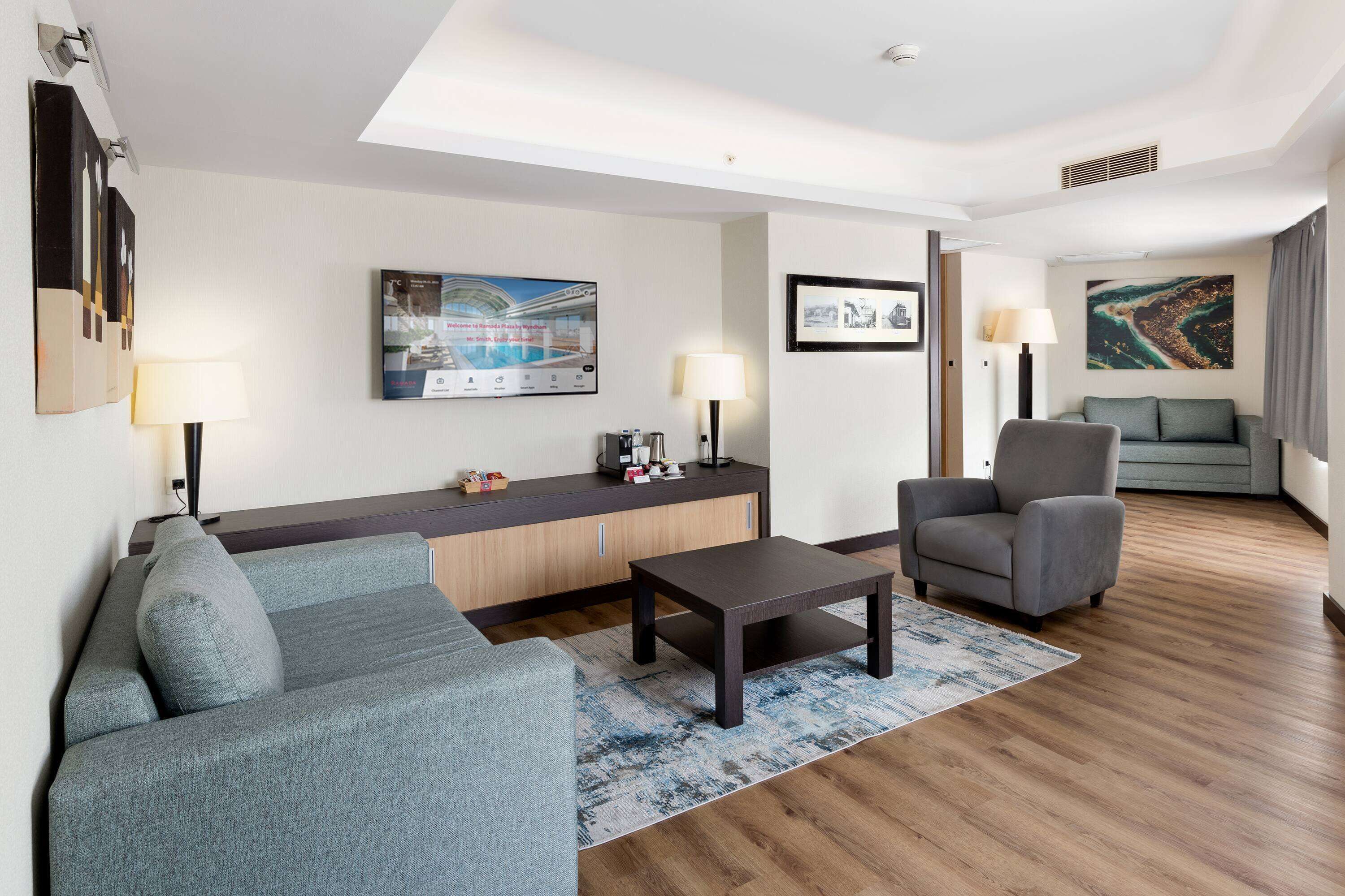 A modern hotel room with a grey sofa, a small table, a grey armchair, a wooden floor, a TV mounted on the wall, and a large window with a view of the sea.