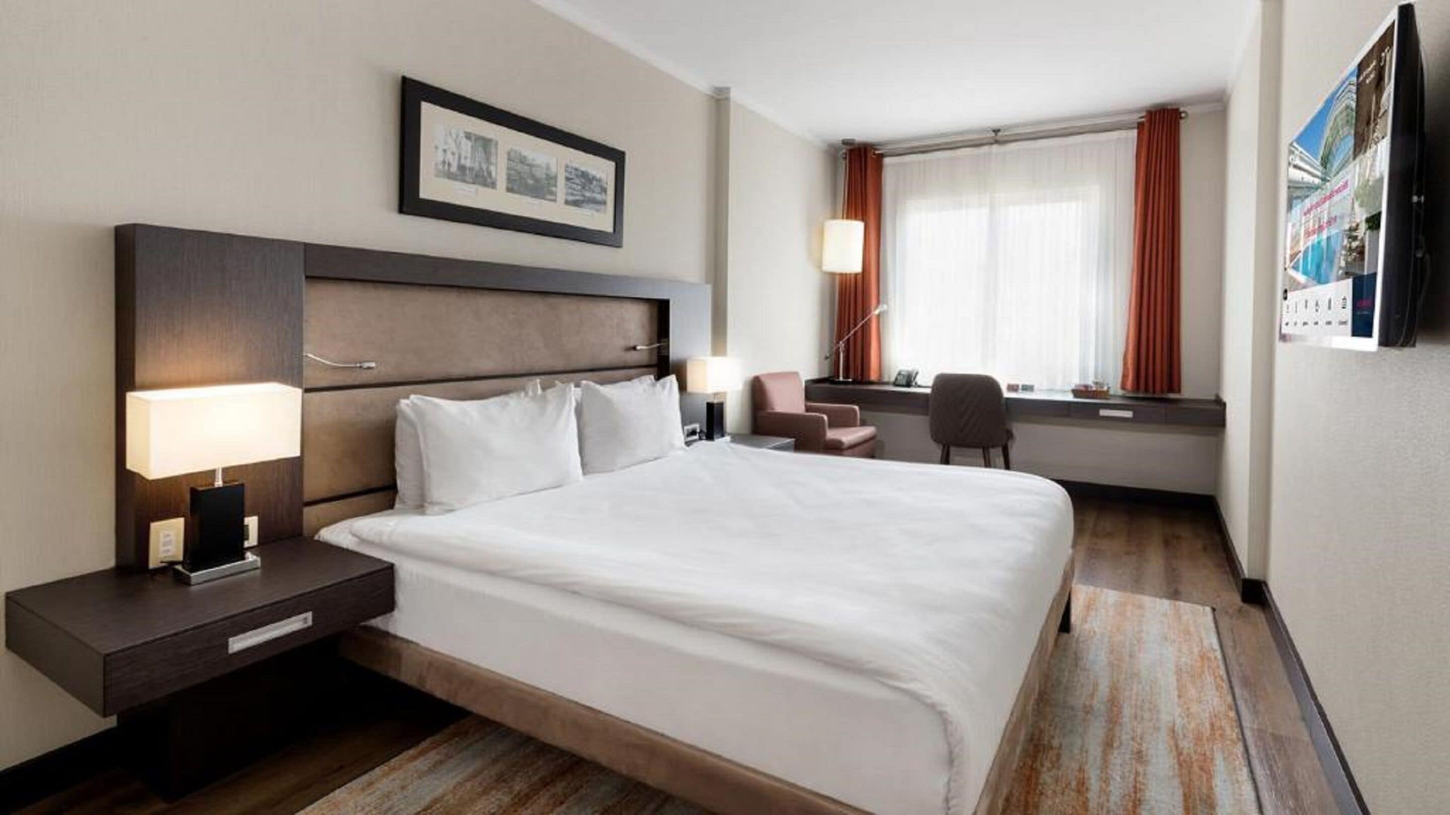 A hotel room with a large bed, bedside tables, a desk, a chair, and a window with curtains.