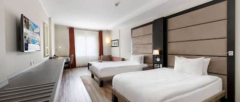 Ein Hotelzimmer mit zwei Betten, einem Schreibtisch, einem Fernseher und einem großen Fenster mit Vorhängen.
