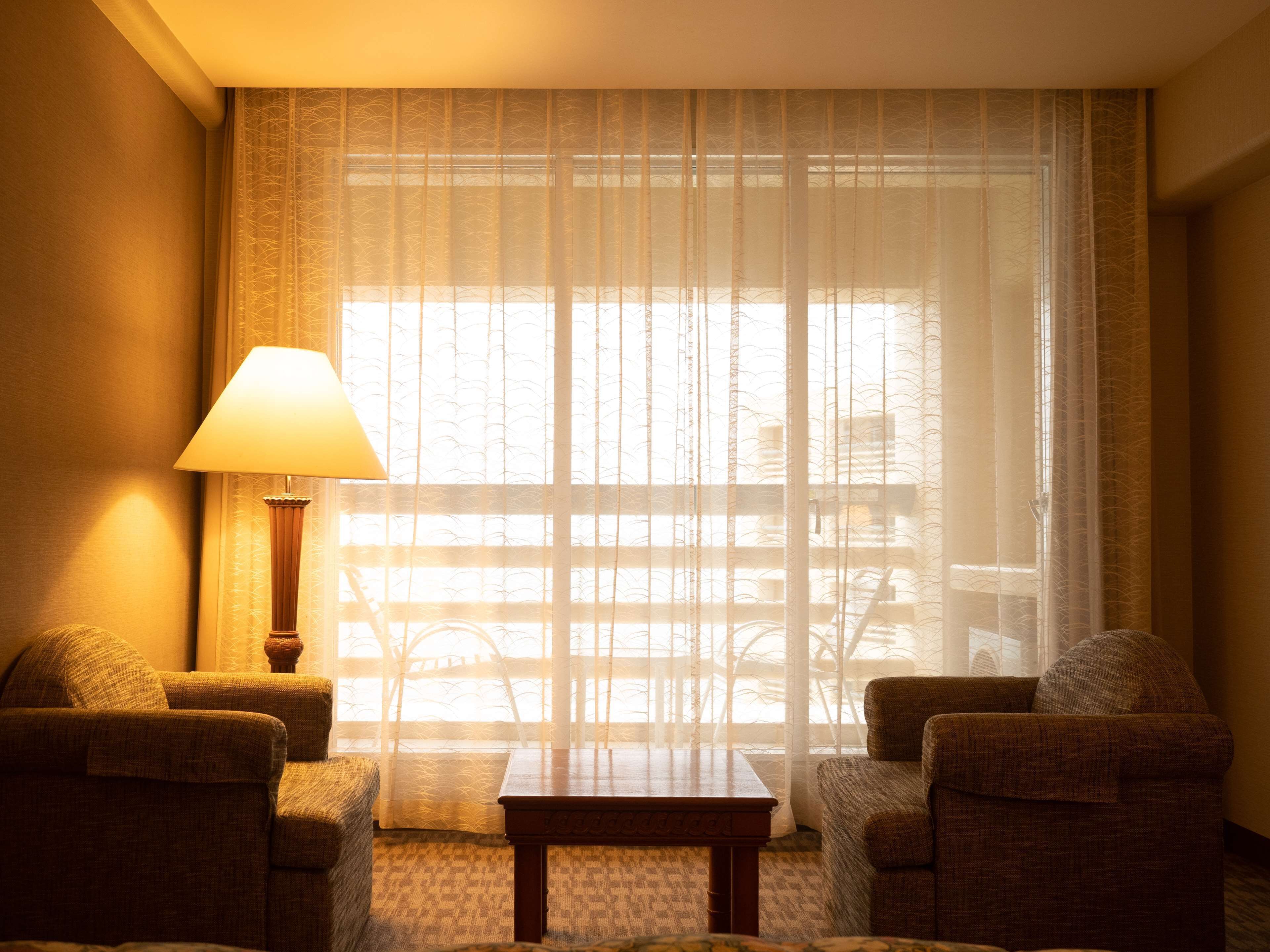 A hotel room with a large window, a lamp, two armchairs, and a small table.