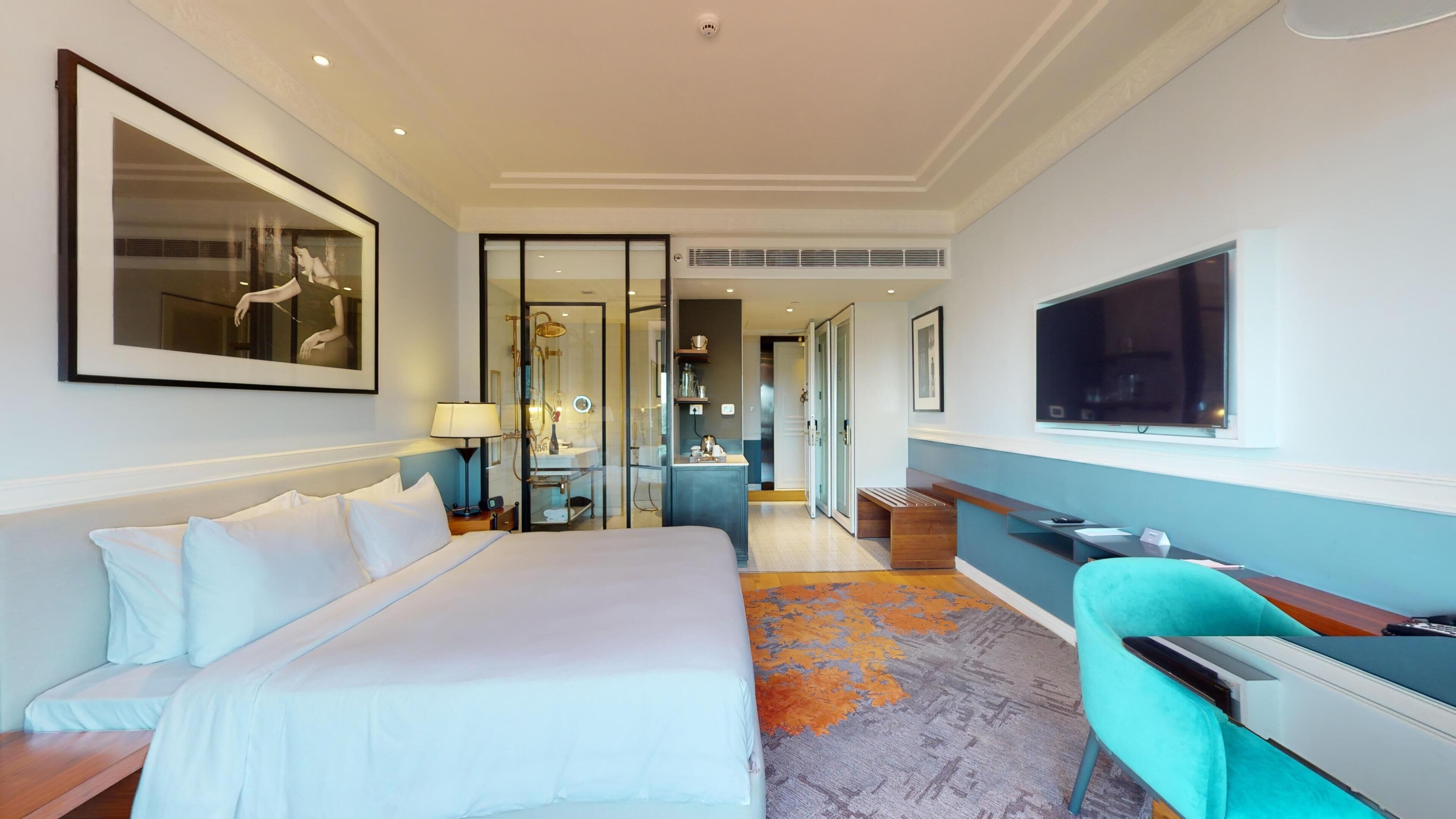 A hotel room with a large bed, a desk with a chair, a TV, and a bathroom visible through a glass door.