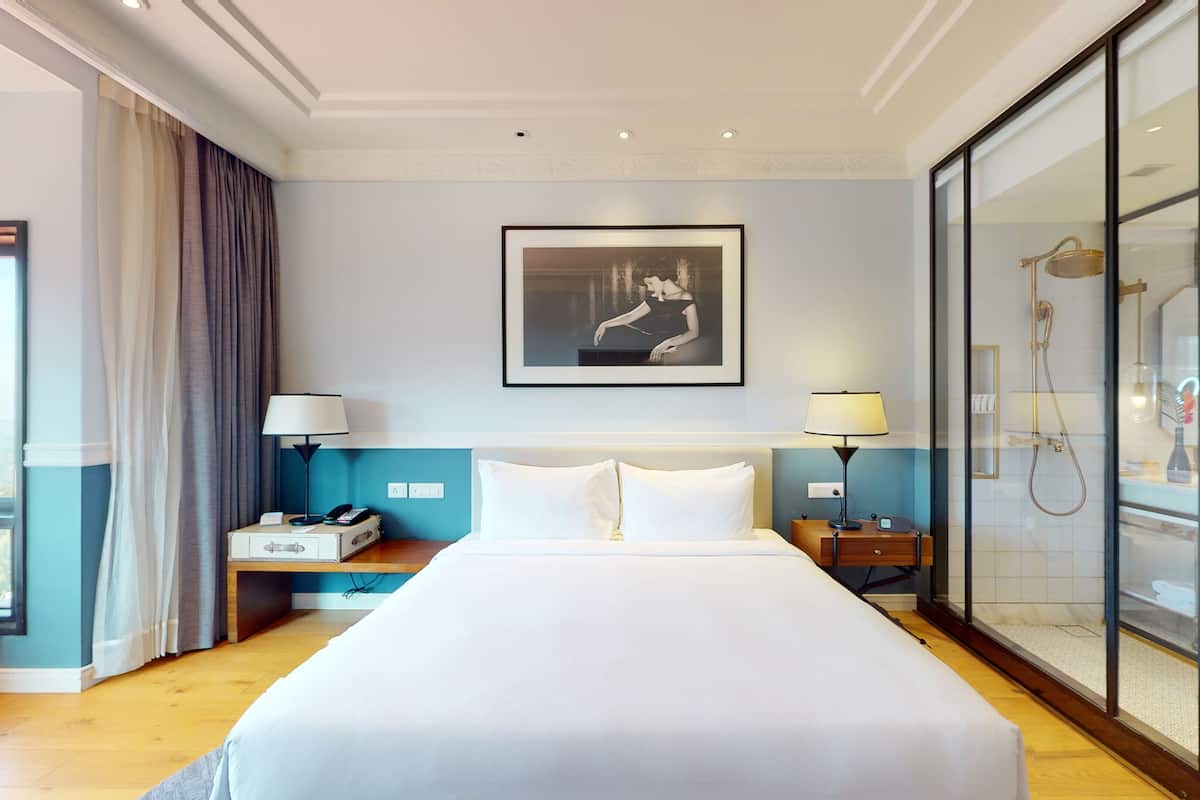 Habitación de hotel con una cama grande, una mesita de noche con lámpara, un atril con teléfono, un cuadro en la pared y una cabina de ducha de vidrio.