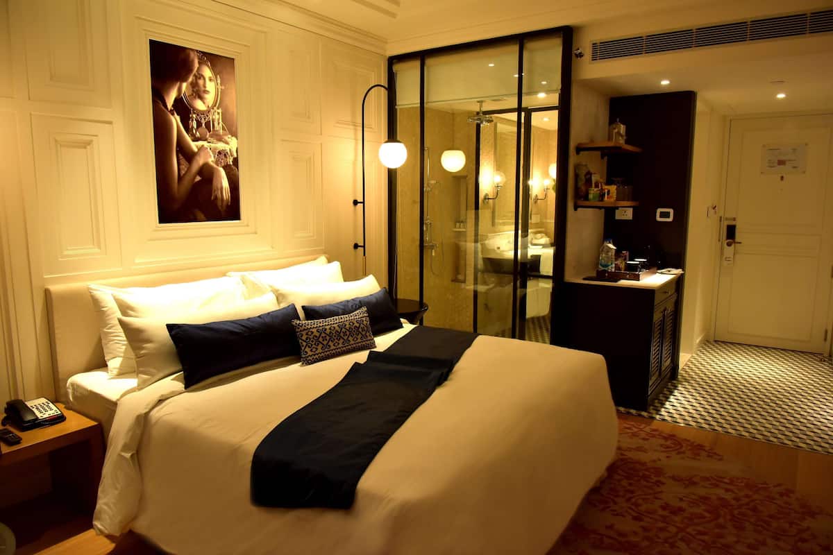 Habitación de hotel con una cama grande, una mesita de noche con un teléfono, zona de ducha con mampara de vidrio y un escritorio pequeño con una lámpara.