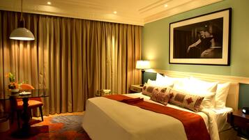 A hotel room with a large bed, a desk with a lamp, a framed picture on the wall, and a curtain-covered window.