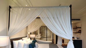 A four-poster canopy bed with white bedding and teal pillows, a bedside table with a lamp, and a nightstand with a remote control.