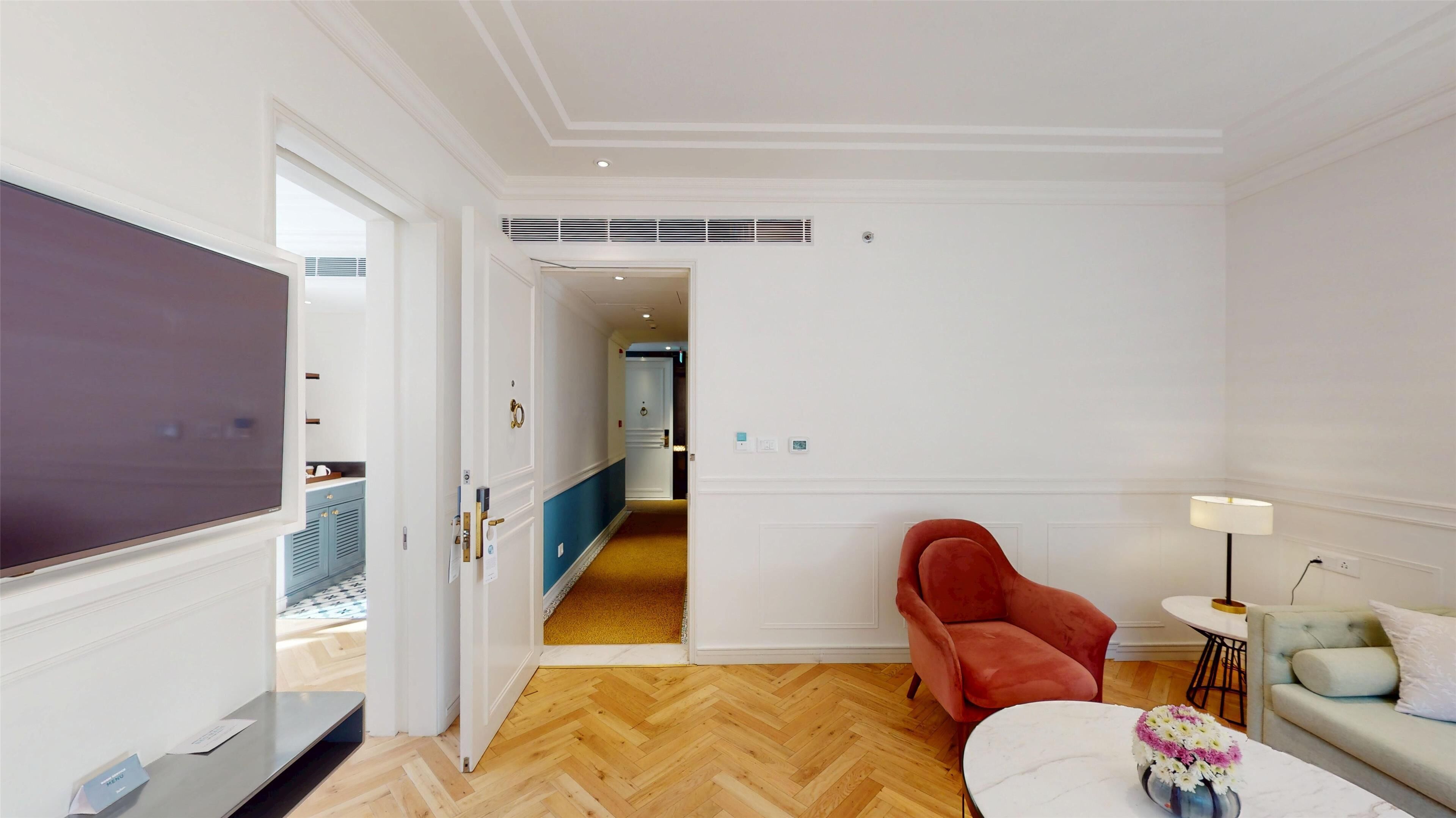A hotel room with a wooden herringbone floor, a red armchair, a green sofa, a white coffee table with flowers, and a flat-screen TV.