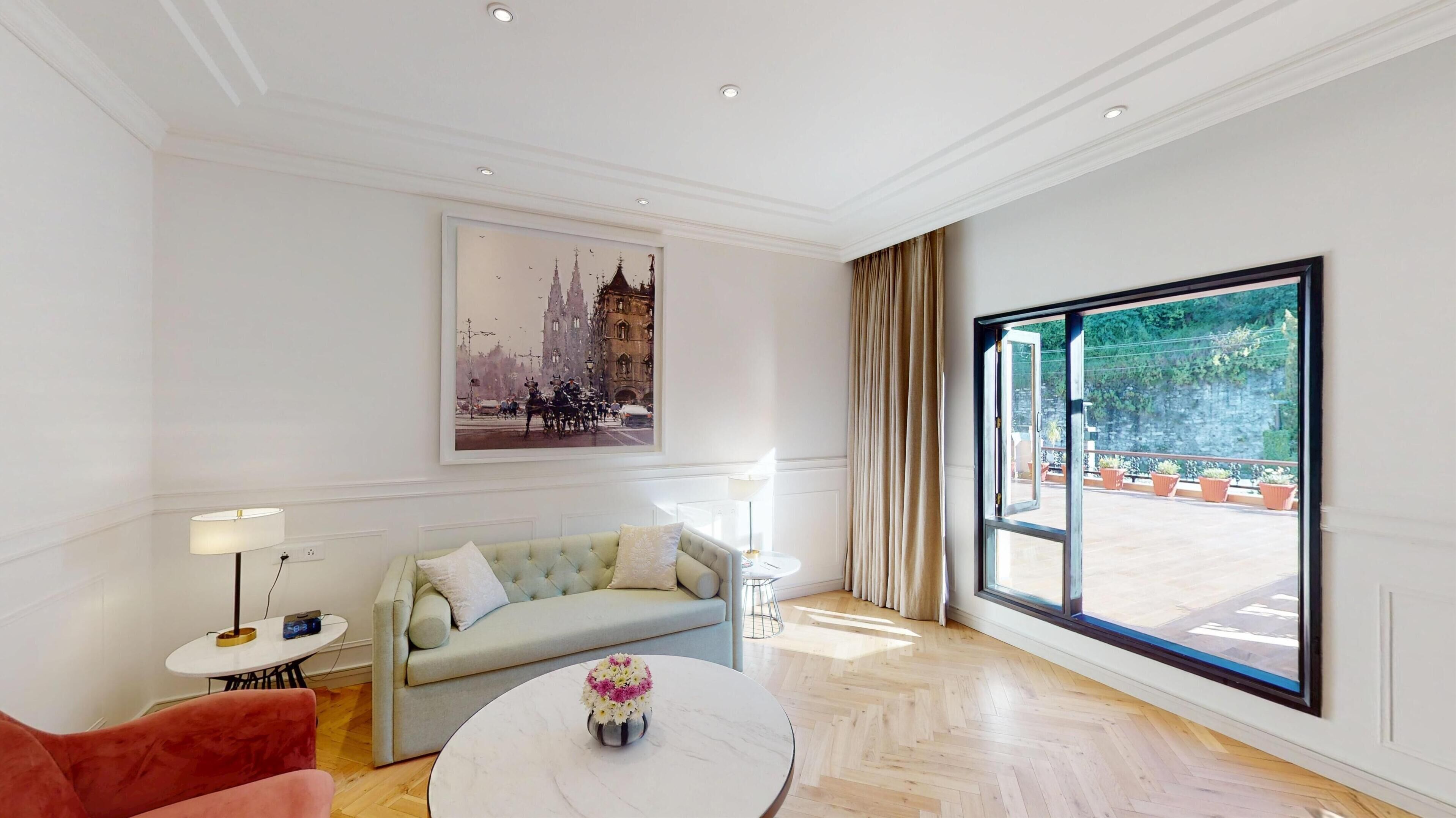Een moderne woonkamer met een bank, een bijzettafeltje met bloemen, een lamp en een groot raam met uitzicht op een balkon.