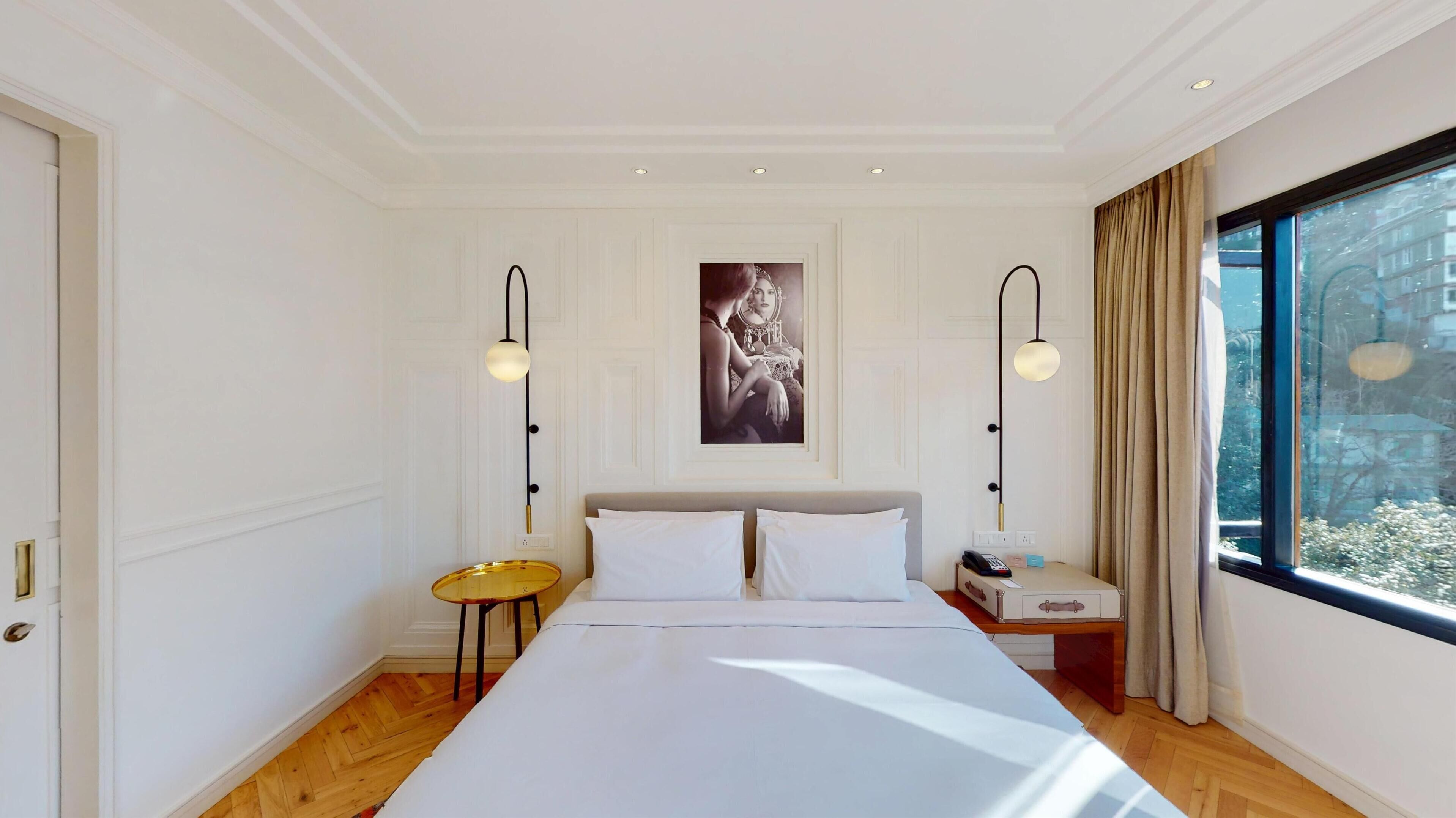 A modern hotel room with a large bed, two bedside tables, a nightstand with a lamp, and a large window with a view of buildings.