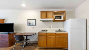 Cocina compacta con armarios de madera, microondas, refrigerador, mesa de comedor con una silla y televisor.