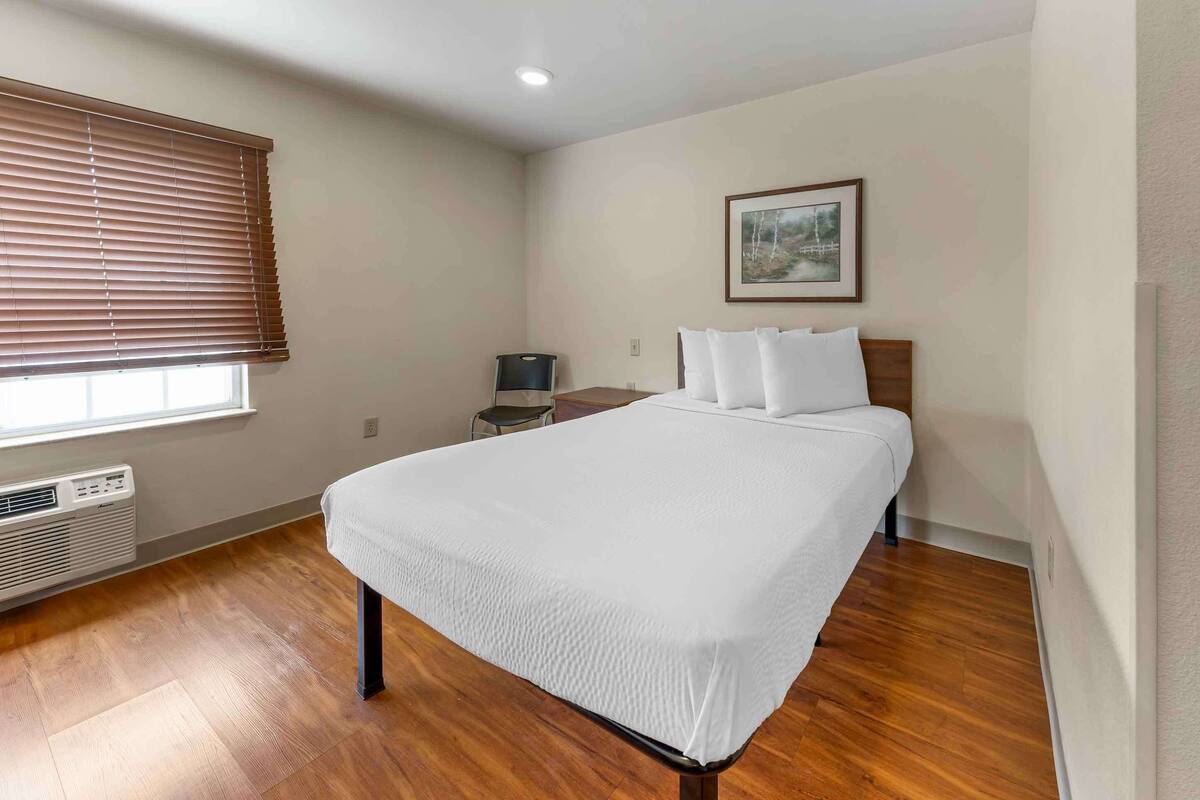 Een slaapkamer met een groot bed, een verwarming, een raam met jaloezieën, een nachtkastje en een lijst met een afbeelding aan de muur.