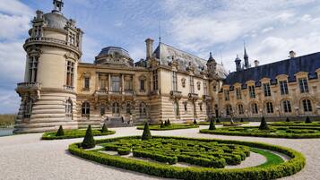A grand historic building with a symmetrical facade, a central courtyard with geometric hedges, and a body of water in the background.