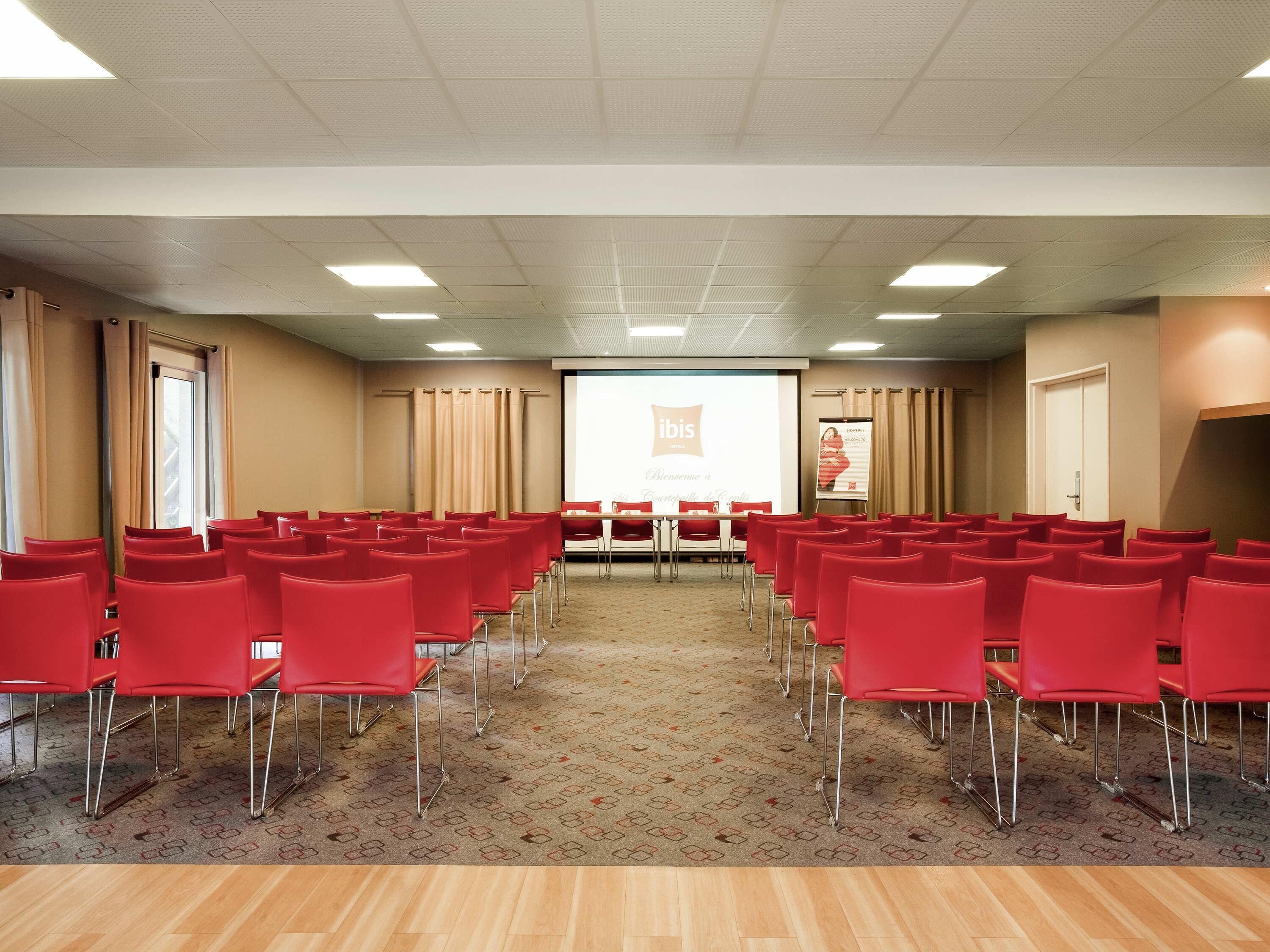 A conference room with rows of red chairs, a carpeted floor, and a projection screen at the front.