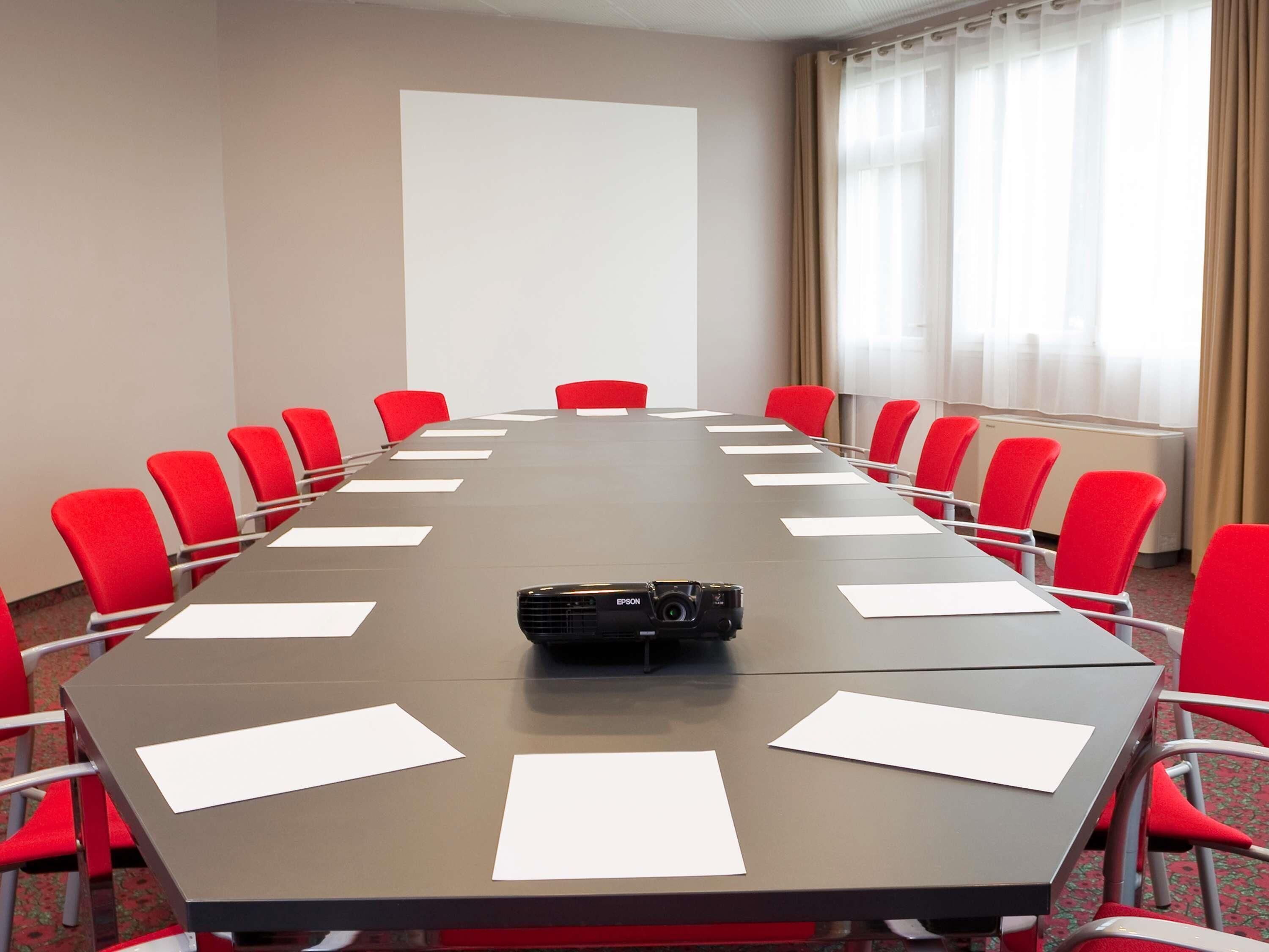 Une salle de conférence avec une longue table, des chaises rouges, un projecteur et un écran vierge.