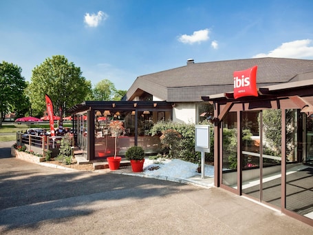 A modern hotel exterior with a covered outdoor seating area, potted plants, and a clear sky.. ibis Senlis