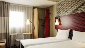 A hotel room with a bed, a desk, a chair, a surfboard, and a wall-mounted lamp. - ibis Senlis (Senlis)