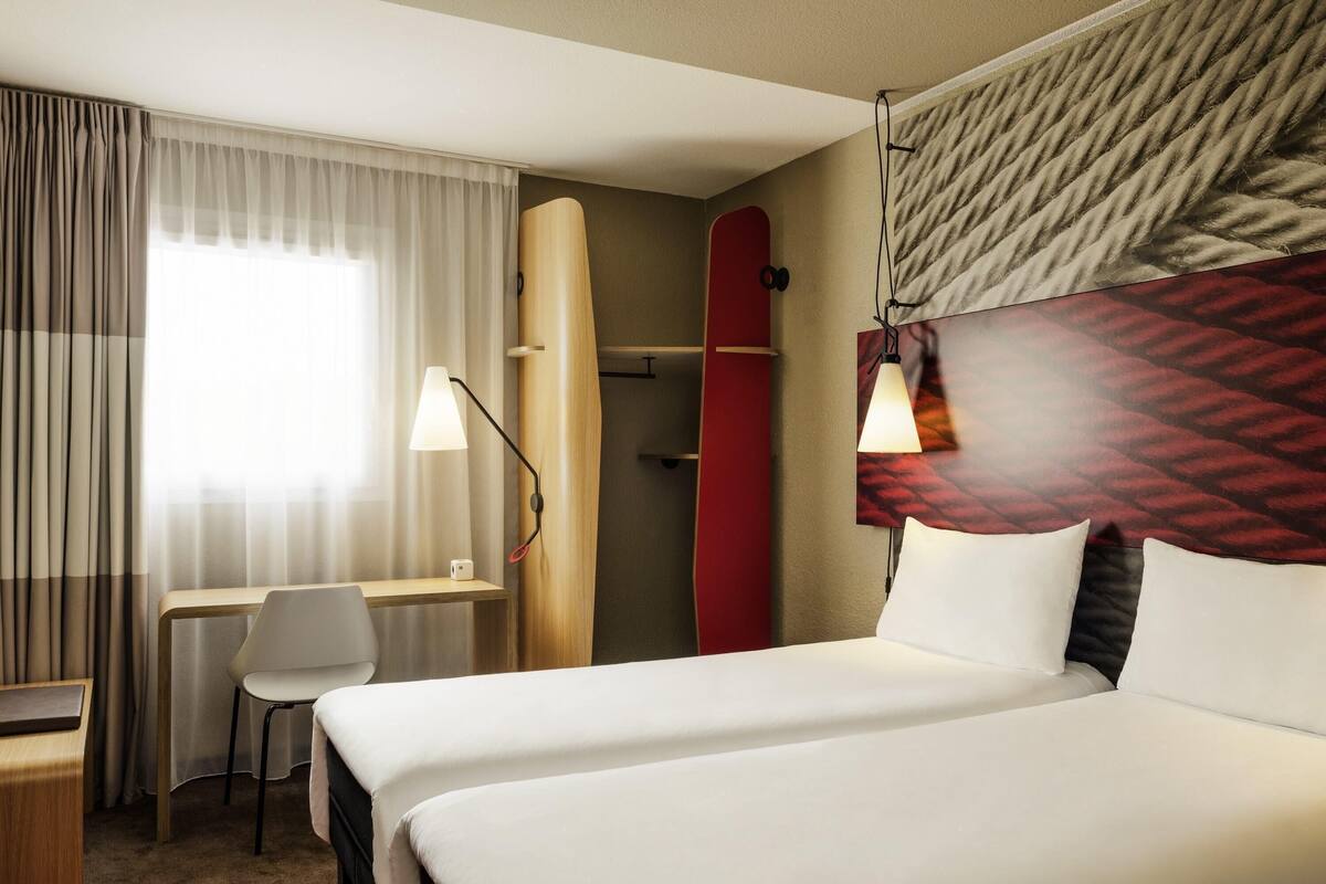 A hotel room with a bed, a desk, a chair, a surfboard, and a wall-mounted lamp.