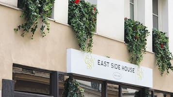 A building with multiple windows, some with green plants and red flowers. The facade has a sign reading 'EAST SIDE HOUSE' with a logo.