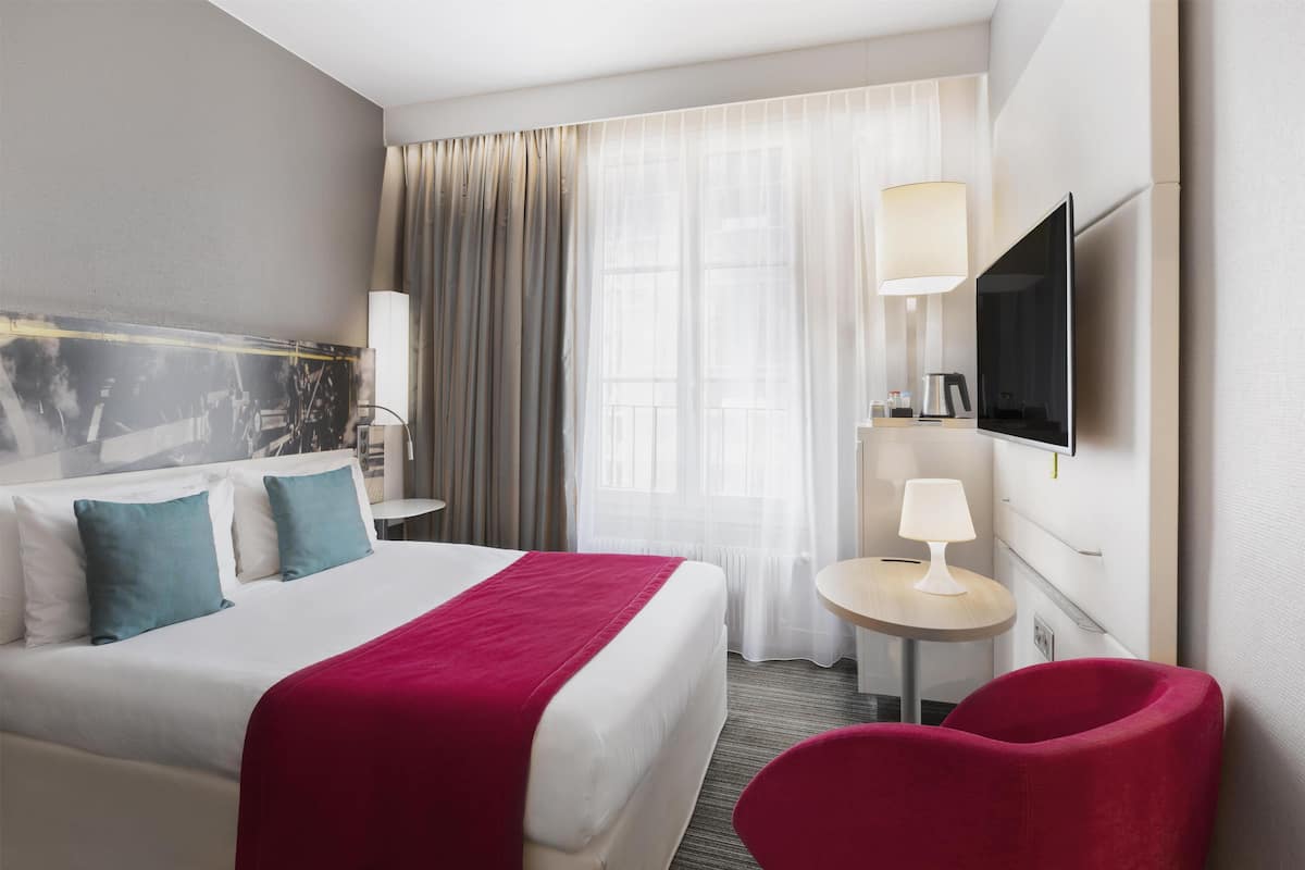 A hotel room with a bed, a TV, a desk, a chair, and a window with curtains.