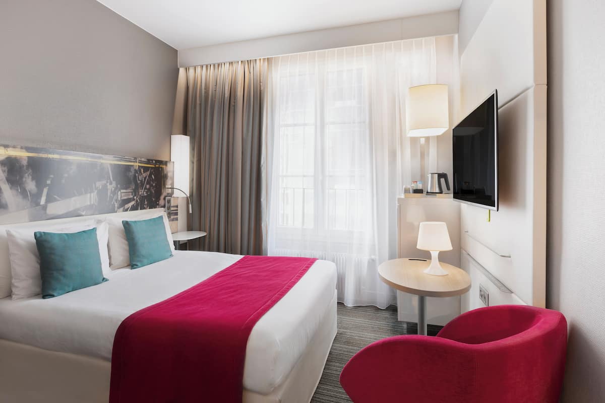 A hotel room with a bed, a TV, a desk, a chair, and a window with curtains.