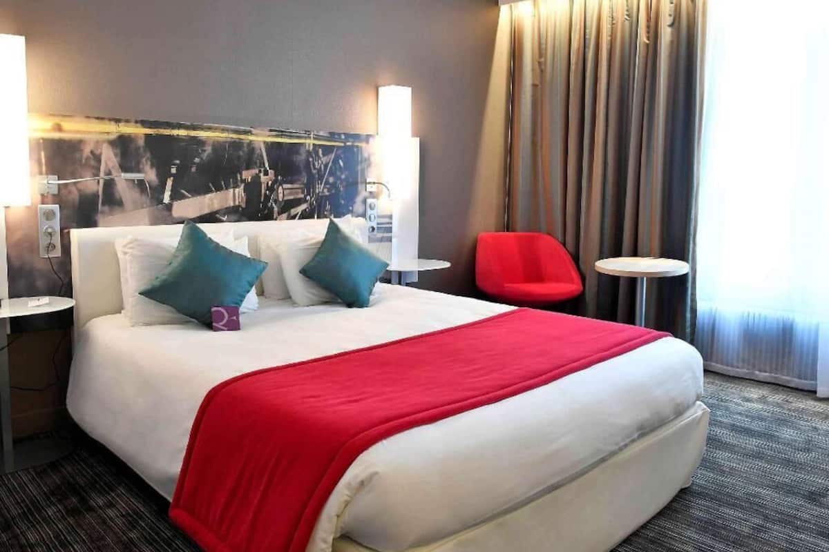 A hotel room with a large bed, two bedside tables, a red chair, a small table, and a view of the outside through curtains.