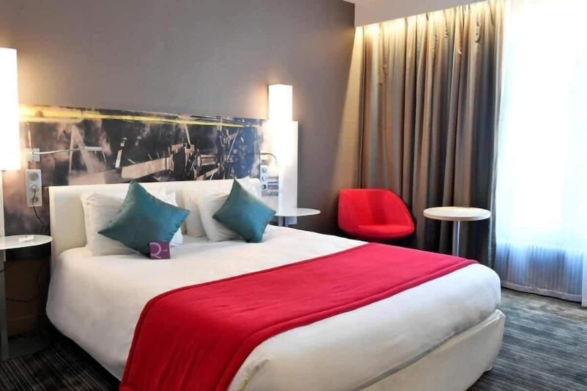 A hotel room with a bed, two bedside tables, a red chair, a small round table, and a large window with curtains.