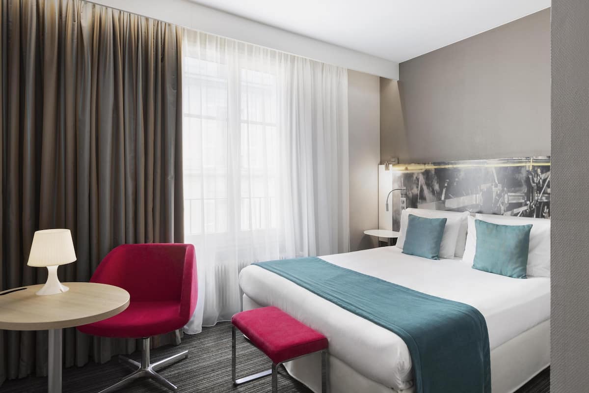 A hotel room with a bed, a red armchair, a small table with a lamp, and a large window with curtains.