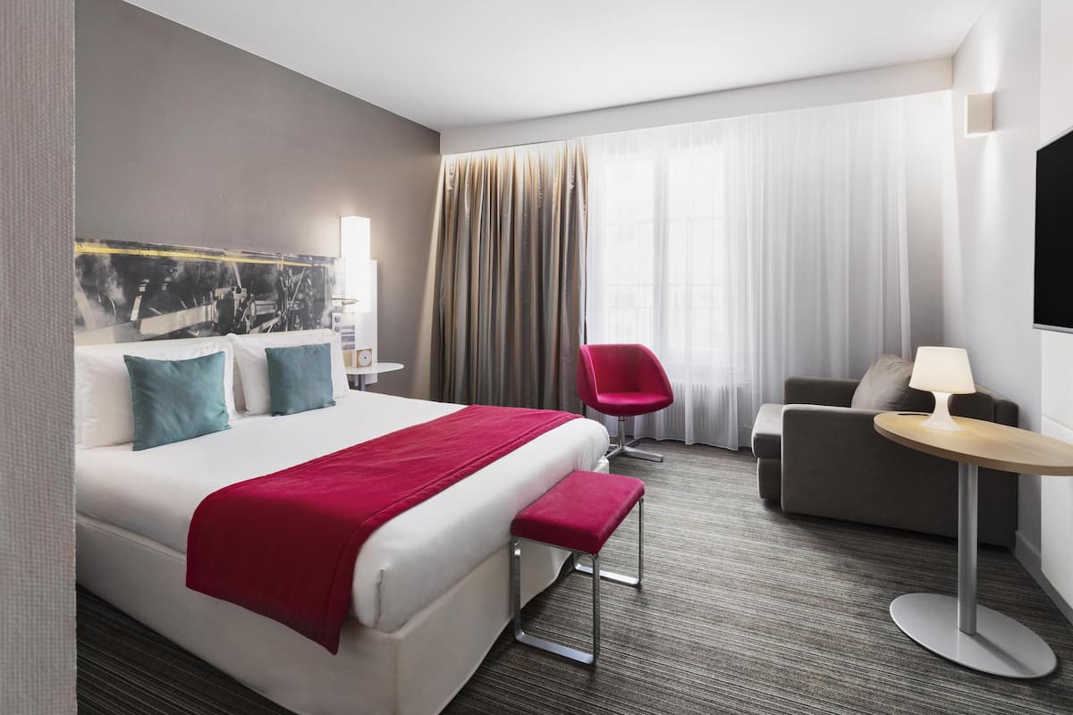 A hotel room with a large bed, a red chair, a sofa, and a small table.