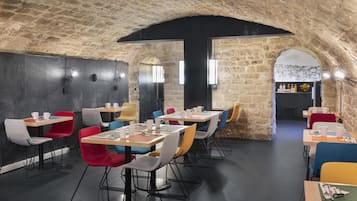 A restaurant with stone walls and modern seating. Tables are arranged in a U-shape, and there are colorful chairs.