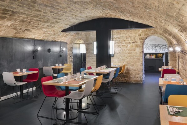 Un restaurante con paredes de piedra y asientos modernos. Las mesas están dispuestas en forma de U, y hay sillas de colores.
