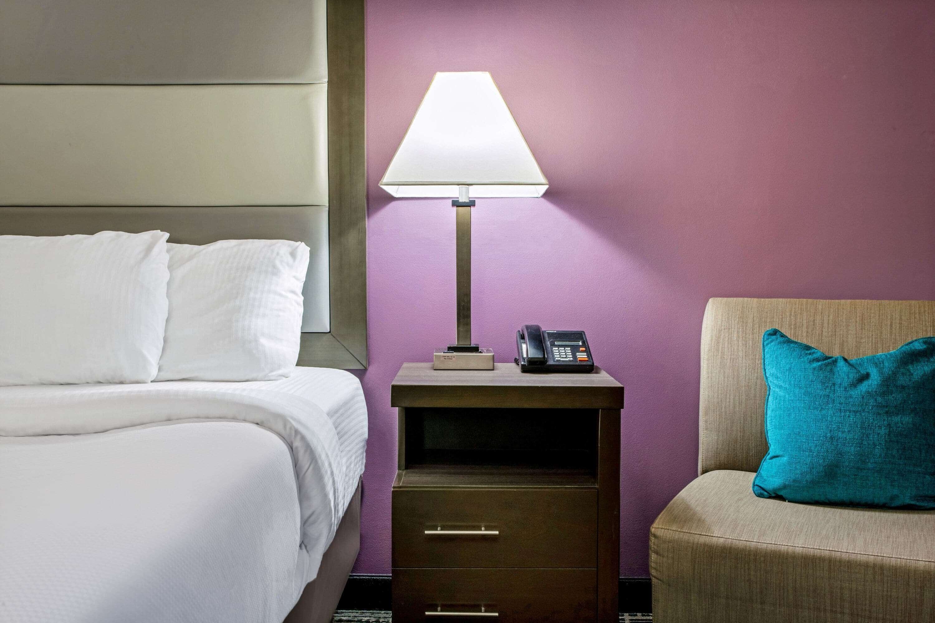 Una habitación de hotel con una cama, una mesita de noche con lámpara, un teléfono y un cojín azul turquesa en una silla.