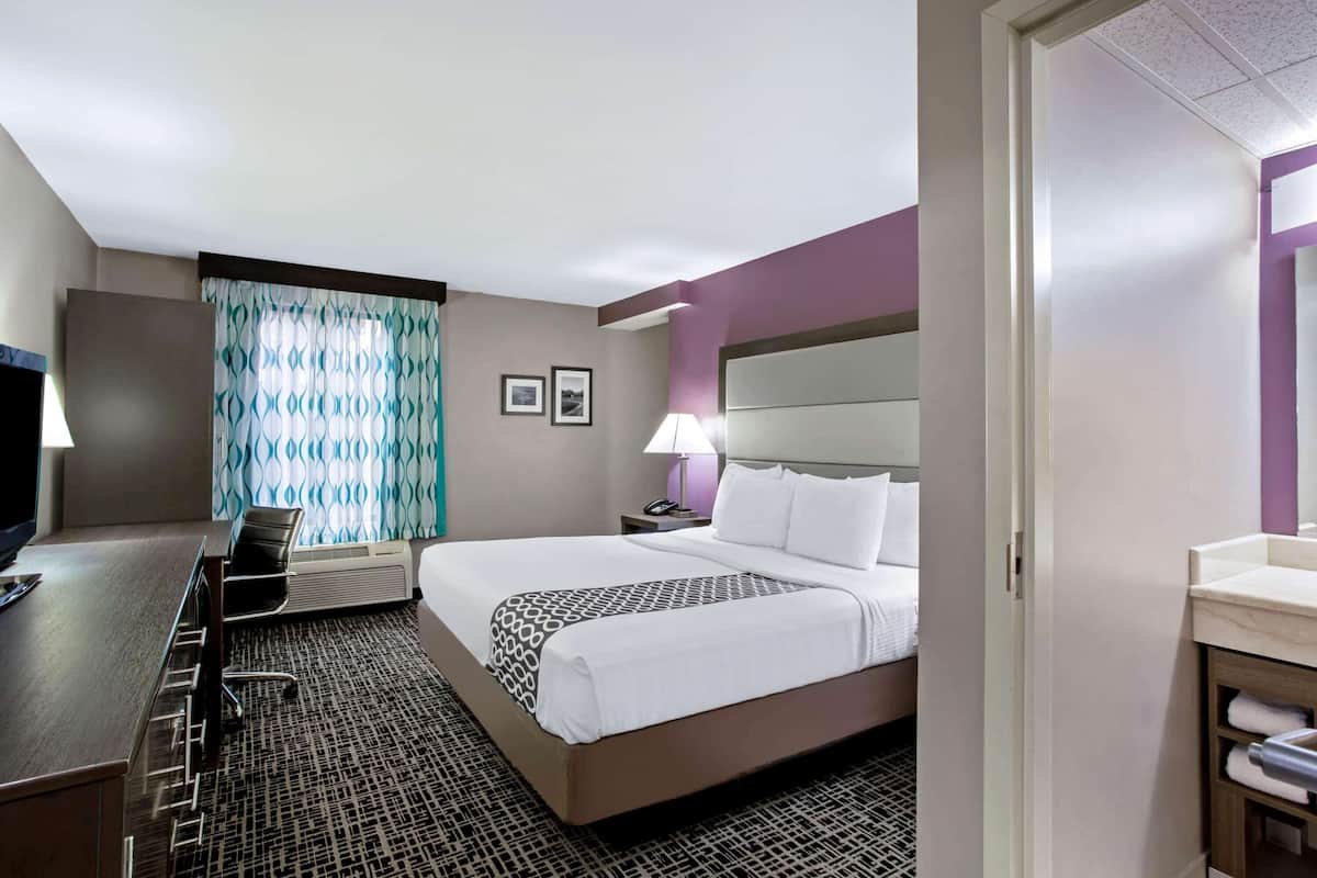 A hotel room with a bed, bedside table, lamp, chair, desk, and a bathroom with a sink and towel rack.