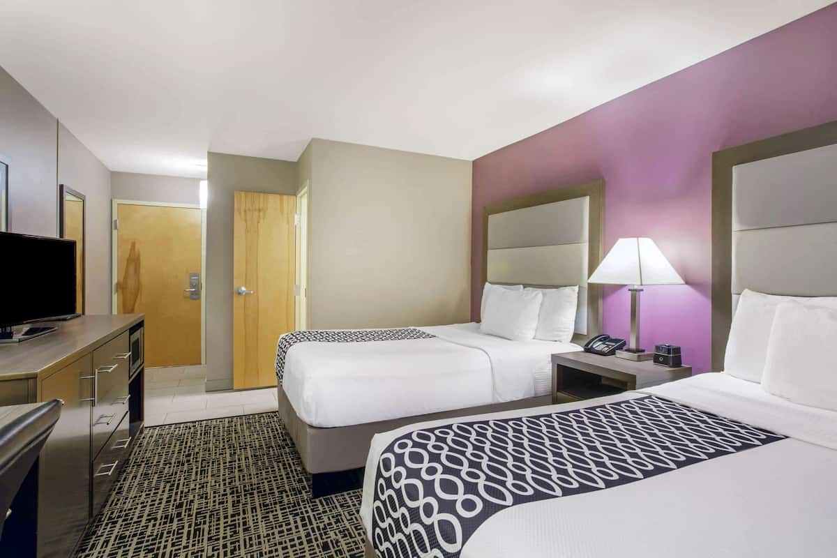 A hotel room with two beds, a desk with a television, a bedside table with a lamp, and a door leading to another room.