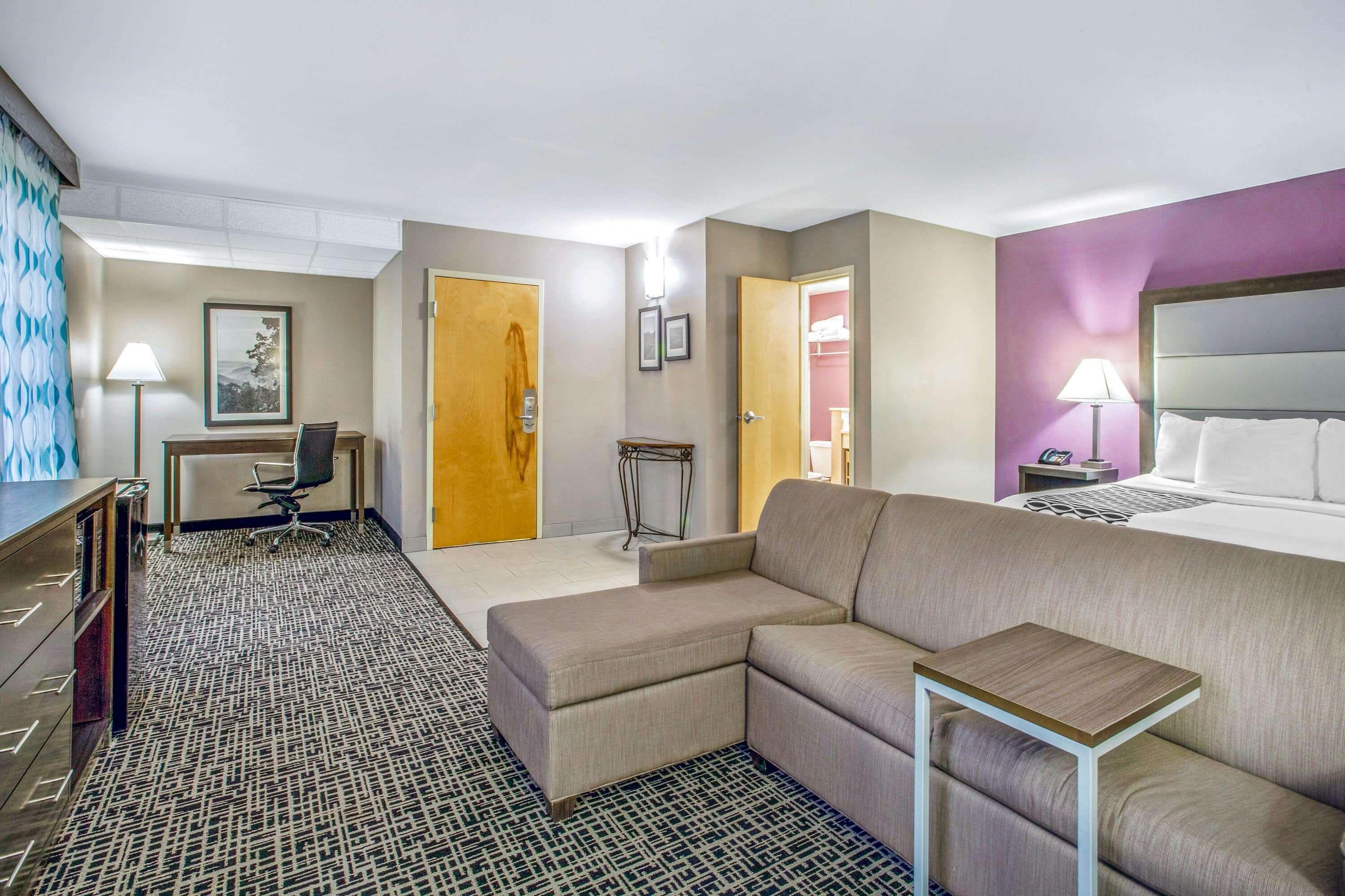 A hotel room with a large bed, a sofa, a desk with a chair, a small table, and a patterned carpet.