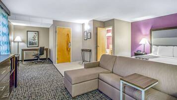 A hotel room with a large bed, a sofa, a desk with a chair, a small table, and a patterned carpet.