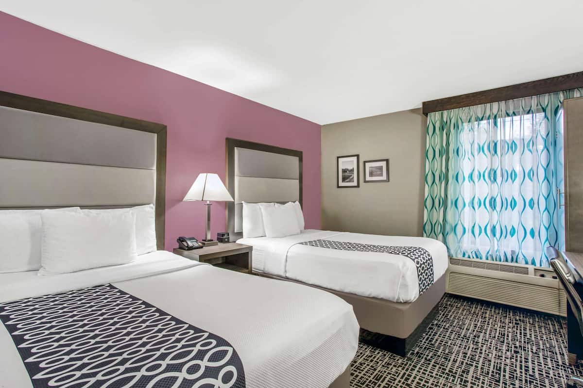 A hotel room with two beds, a nightstand with a lamp, a window with patterned curtains, and a wall-mounted phone.