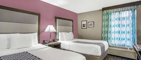 A hotel room with two beds, a nightstand with a lamp, a window with patterned curtains, and a wall-mounted phone.