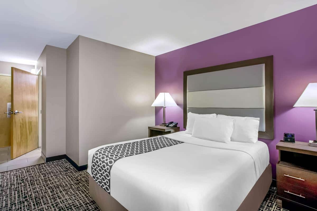 A hotel room with a large bed, a bedside table with a lamp, a nightstand with a phone, and a wooden door.