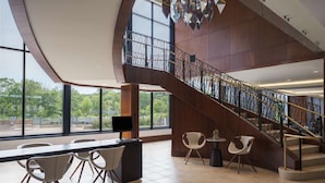 A spacious lobby with a modern staircase, large windows, and a seating area with chairs and a table.