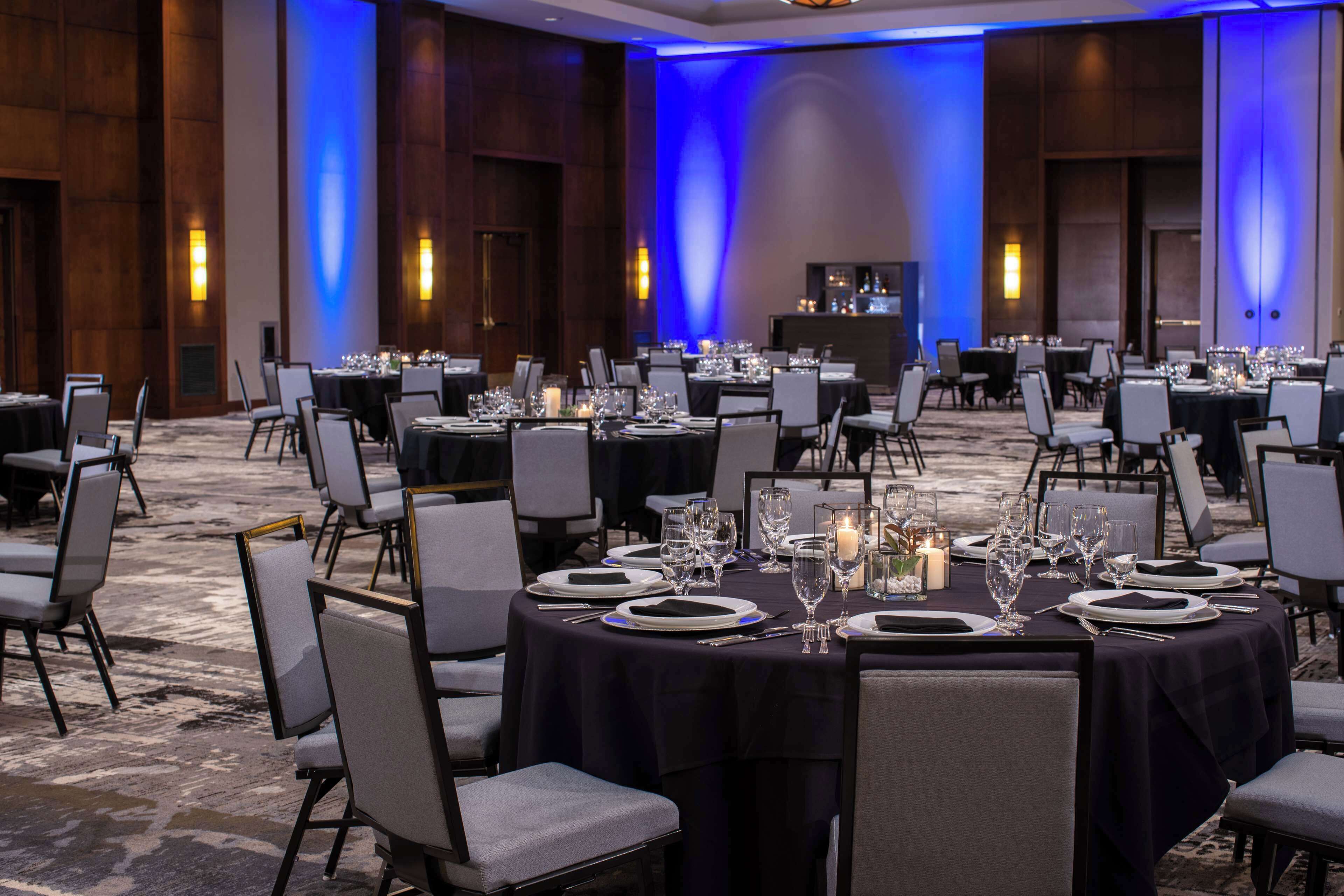 Un salón de banquetes con mesas redondas dispuestas para el servicio de comidas, equipado con sillas, platos, cubiertos y velas.